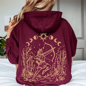 May include: A maroon hooded sweatshirt featuring a gold-colored graphic on the back. The design depicts an archer aiming a bow within a forest setting, surrounded by a star and a series of moon phases arranged in an arc above the figure.