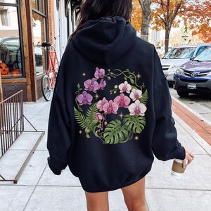 Puede incluir: Una sudadera negra con capucha que presenta un gran gr&aacute;fico circular en la espalda. El dise&ntilde;o muestra flores de orqu&iacute;dea moradas y blancas, hojas de monstera verdes y frondas de helecho dispuestas en corona, con peque&ntilde;os iconos de estrellas doradas.