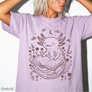 May include: Orchid-colored t-shirt featuring a brown axolotl reading a book, surrounded by floral and celestial elements. The design includes a crescent moon and butterflies. The word "Orchid" is printed at the bottom.