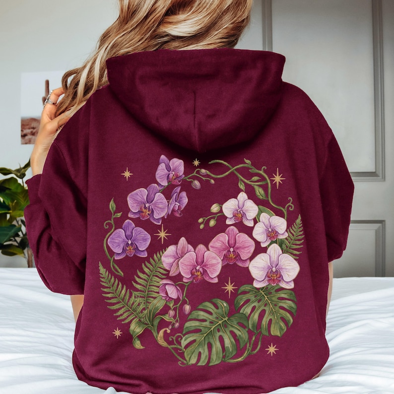 Puede incluir: Una sudadera con capucha color granate que presenta un gran gr&aacute;fico en la espalda de orqu&iacute;deas moradas y blancas, frondas de helecho verdes y hojas de monstera dispuestas en c&iacute;rculo, acentuado con peque&ntilde;as estrellas doradas de cuatro puntas.
