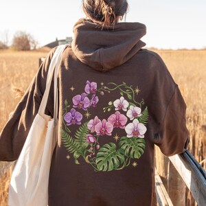 Puede incluir: Una sudadera con capucha de color marr&oacute;n oscuro que presenta un gran gr&aacute;fico en la espalda de orqu&iacute;deas moradas y blancas, frondas de helecho verdes y hojas de monstera dispuestas en c&iacute;rculo, con peque&ntilde;os iconos de estrellas doradas y un bolso blanco.