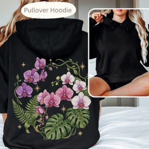 Puede incluir: Una sudadera con capucha negra que presenta un gran gr&aacute;fico en la espalda de orqu&iacute;deas moradas y blancas, frondas de helecho verdes y hojas de monstera acentuadas con peque&ntilde;as estrellas doradas. El frente es negro liso con capucha.