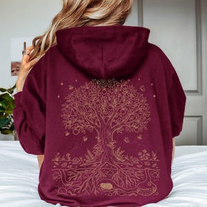 Op de afbeelding: Bordeaux hoodie met een goudkleurig levensboomontwerp op de rug. De boom heeft fruit, bloemen en wortels, met insecten en kleine dieren. De hoodie heeft een capuchon en lange mouwen en is gemaakt van een zacht materiaal.