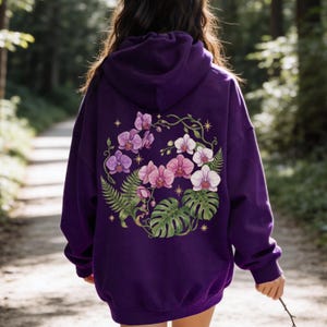 Puede incluir: Una sudadera con capucha de color morado oscuro que presenta un gran gr&aacute;fico en la espalda de orqu&iacute;deas rosas y blancas, frondas de helecho verdes y hojas de monstera dispuestas en c&iacute;rculo, con peque&ntilde;os s&iacute;mbolos de estrellas doradas de cuatro puntas.