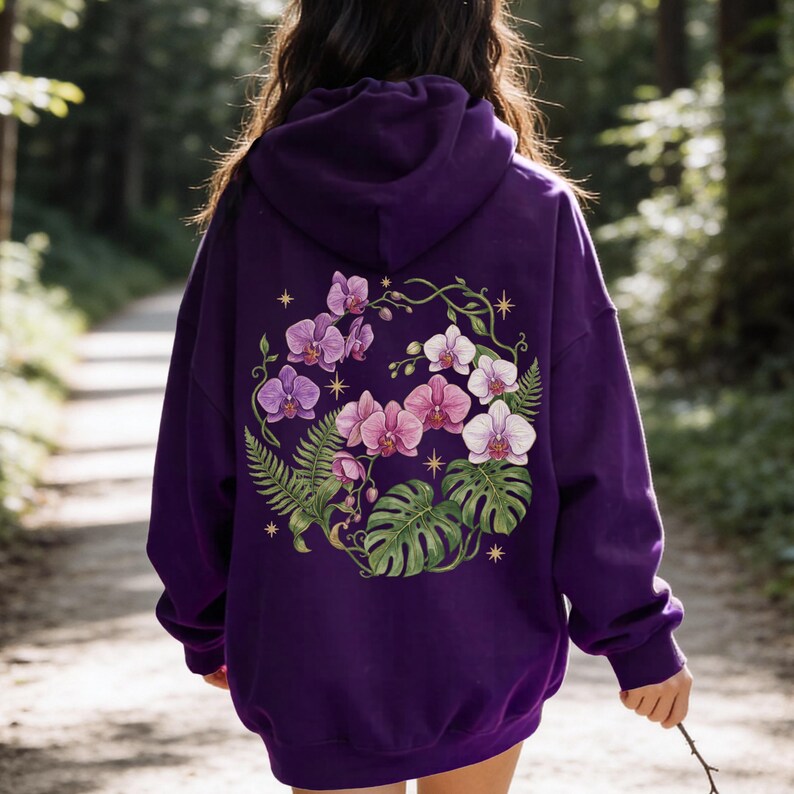 Puede incluir: Una sudadera con capucha de color morado oscuro que presenta un gran gr&aacute;fico en la espalda de orqu&iacute;deas rosas y blancas, frondas de helecho verdes y hojas de monstera dispuestas en c&iacute;rculo, con peque&ntilde;os s&iacute;mbolos de estrellas doradas de cuatro puntas.