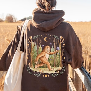 May include: Brown hoodie featuring a whimsical illustration of a Bigfoot character in a forest scene with butterflies and mushrooms. The design includes a crescent moon and stars. The hoodie has a drawstring hood and long sleeves.