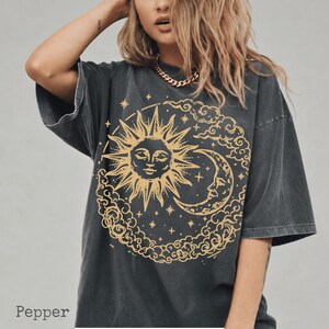 Könnte beinhalten: Ein anthrazitfarbenes Oversize-T-Shirt mit einem großen goldenen Grafikdruck von Sonne und Mondsichel, umgeben von Wolken und Sternen. Das Wort Pepper ist in der unteren linken Ecke des Bildes in einer einfachen dunkelgrauen Schrift gedruckt.