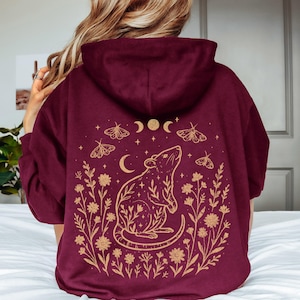 May include: A maroon hooded sweatshirt featuring a gold-colored graphic on the back. The design depicts a rat surrounded by wildflowers, moths, stars, and moon phases. The hoodie has long sleeves and a hood, showcasing a detailed botanical illustration.