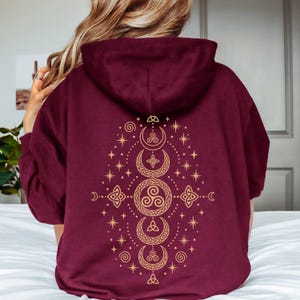 Op de afbeelding: Een dieprode hoodie met een grote, gedetailleerde goudkleurige afbeelding op de achterkant. Het ontwerp toont een verticale opstelling van hemelse manen, sterren en Keltisch geïnspireerde knooppatronen, inclusief een centrale triskelion.
