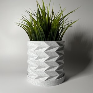 May include: A white geometric planter with a textured surface holds a green artificial plant. The planter has a matching saucer.