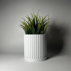 May include: A white planter with a ribbed design and a green artificial plant. The planter has a textured surface.