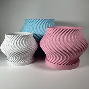 Könnte beinhalten: Drei Keramik-Blumentöpfe in verschiedenen Größen mit einem wellenförmigen Design. Die Töpfe sind weiß, hellblau und rosa.