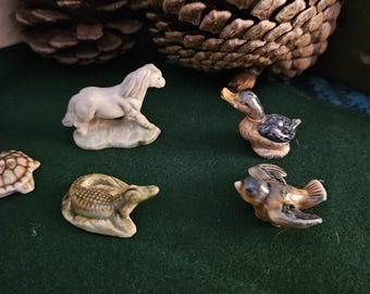 Może przedstawiać: Kolekcja małych ceramicznych figurek zwierząt, w tym białego konia, żółwia, kaczki, ryby i aligatora. Figurki umieszczone są na ciemnozielonej powierzchni, z szyszkami w tle.