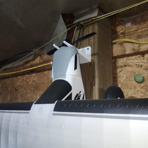 Könnte beinhalten: Ein teilweise montiertes Modellflugzeug mit einem weißen und schwarzen Flügel und einem grau-weißen Rumpf. Der Propeller ist schwarz und das Modell ist an einer Holzwand befestigt.
