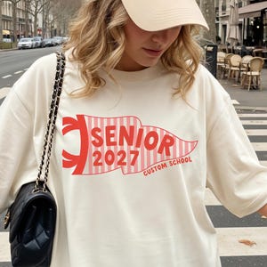 May include: An ivory t-shirt featuring a red and white striped pennant graphic with the text SENIOR 2027 CUSTOM SCHOOL, paired with a beige baseball cap with a small dark logo and a black shoulder bag with a gold chain strap.
