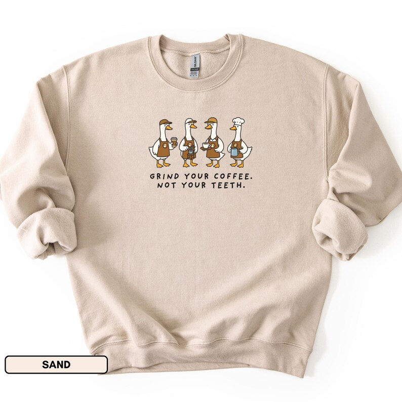 May include: A sand-coloured crew neck sweatshirt. The graphic shows four cartoon geese wearing aprons and hats, holding coffee cups. The text "GRIND YOUR COFFEE. NOT YOUR TEETH." is printed below the geese.
