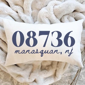 May include: Cream-colored rectangular pillow with the numbers "08736" in navy blue, and the words "manasquan, nj" in a script font. The pillow is resting on a fluffy, cream-colored blanket.
