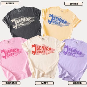 May include: Five short-sleeved crew neck t-shirts in pepper, butter, blossom, ivory, and orchid colors. Each shirt features a graphic of a striped pennant flag with the text SENIOR 2027 CUSTOM SCHOOL printed in a bold, sans-serif font across the center.