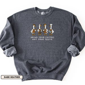 May include: Dark grey sweatshirt featuring a graphic of four geese, each holding a coffee cup. The geese wear aprons and hats. The text reads "GRIND YOUR COFFEE. NOT YOUR TEETH." below the geese.