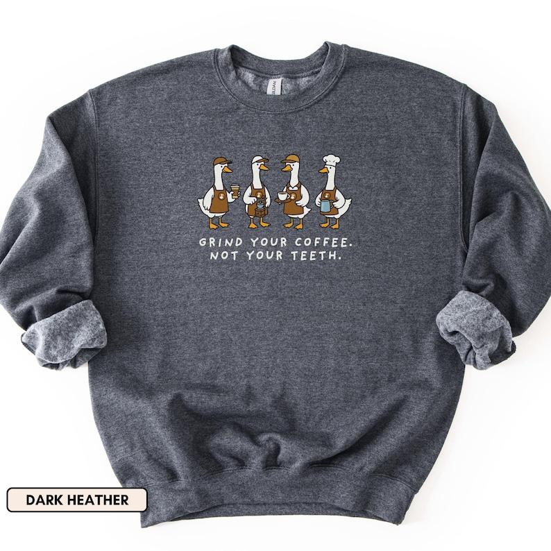 May include: Dark grey sweatshirt featuring a graphic of four geese, each holding a coffee cup. The geese wear aprons and hats. The text reads "GRIND YOUR COFFEE. NOT YOUR TEETH." below the geese.