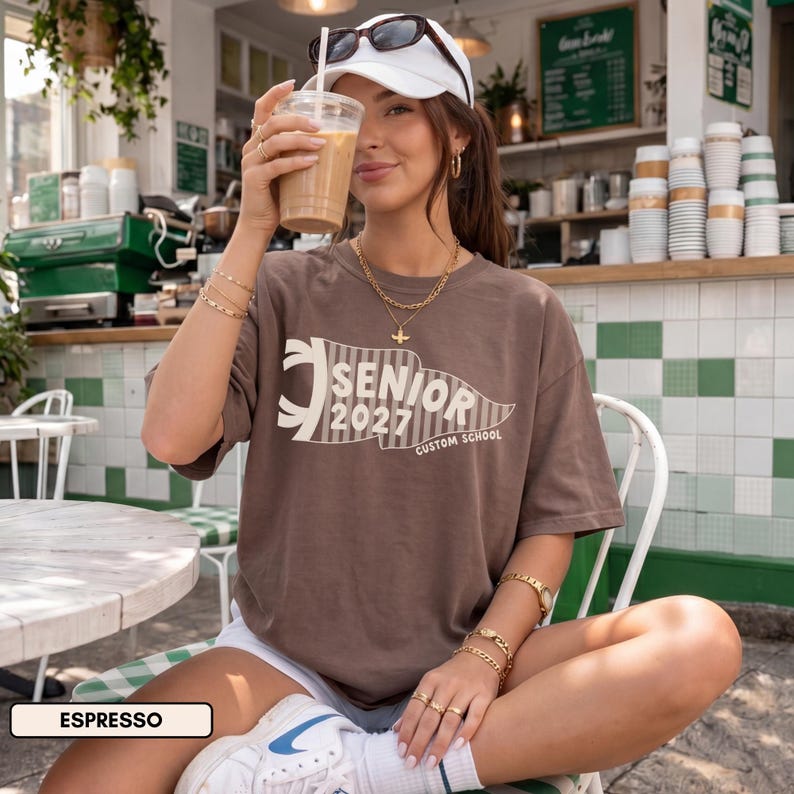 May include: A brown short-sleeve t-shirt featuring a cream-colored pennant graphic with the text SENIOR 2027 CUSTOM SCHOOL. The shirt has a crew neckline and a relaxed fit, made from a solid-colored cotton fabric in a deep espresso brown shade.
