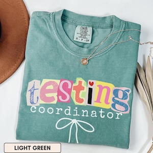 May include: Light green t-shirt with the words "testing coordinator" in colorful, cut-out style letters. A small heart is above the letter "i". A silver necklace with a pendant is on the shirt. The shirt is labeled "Comfort Colors".