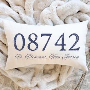 May include: Cream-colored rectangular pillow with the navy blue text "08742" and "Pt. Pleasant, New Jersey". The pillow is resting on a soft, white, textured blanket.