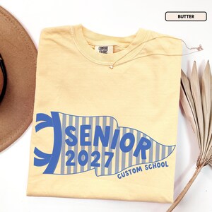 May include: A butter yellow short-sleeve t-shirt with a Comfort Colors label at the neck. The front features a blue and white striped pennant graphic with the text SENIOR 2027 and CUSTOM SCHOOL printed in bold blue lettering across the center.