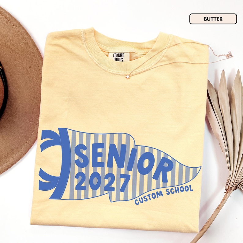 May include: A butter yellow short-sleeve t-shirt with a Comfort Colors label at the neck. The front features a blue and white striped pennant graphic with the text SENIOR 2027 and CUSTOM SCHOOL printed in bold blue lettering across the center.