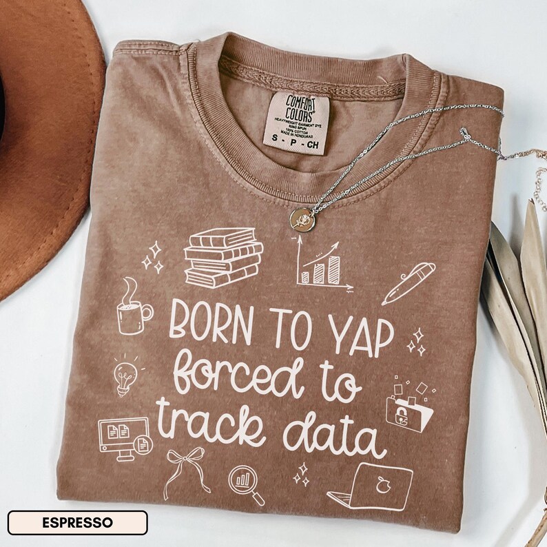 May include: Espresso-colored Comfort Colors t-shirt with the phrase "BORN TO YAP forced to track data" in white. The shirt features graphic icons of books, a coffee cup, a lightbulb, and a laptop. A silver necklace with a pendant is draped across the shirt.