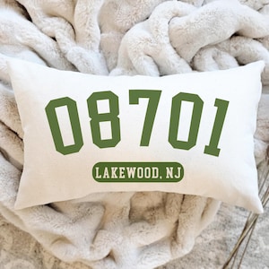 May include: Cream-colored rectangular pillow with the green numbers "08701" and the text "LAKEWOOD, NJ". The pillow is resting on a soft, cream-colored blanket. A decorative accent for a home.