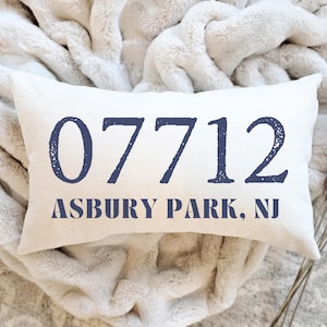 May include: Cream-colored rectangular pillow with the text "07712 ASBURY PARK, NJ" in navy blue. The pillow is resting on a soft, white, textured blanket. The pillow is a decorative item.