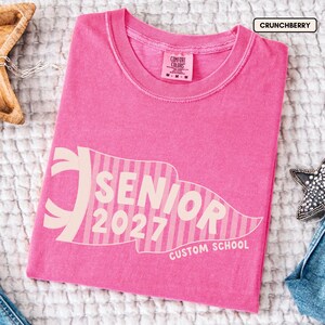 May include: A bright pink short-sleeve t-shirt featuring a large, light pink striped pennant graphic on the front. The text inside the pennant reads SENIOR 2027, with CUSTOM SCHOOL printed below. The shirt has a Comfort Colors label in the neck.