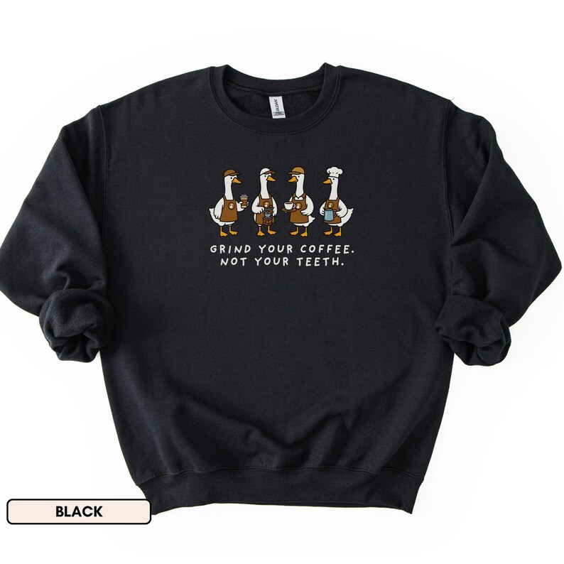 May include: Black sweatshirt with a graphic of four geese in barista outfits, holding coffee cups. The text "GRIND YOUR COFFEE. NOT YOUR TEETH." is printed below. The sweatshirt has a crew neck and long sleeves, perfect for cooler weather.