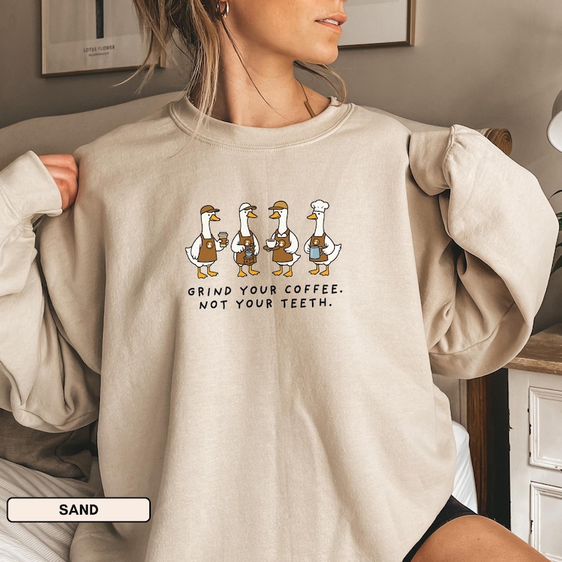 May include: A sand-coloured sweatshirt with a crew neck and long sleeves. It features a cartoon illustration of four geese, each holding coffee-related items, dressed as baristas. The text "GRIND YOUR COFFEE. NOT YOUR TEETH." is printed below the geese.