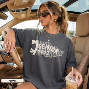 May include: A charcoal gray short-sleeve t-shirt featuring a cream-colored pennant graphic with vertical stripes. The text inside the pennant reads SENIOR 2027 and CUSTOM SCHOOL in a bold, sans-serif font. The shirt has a crew neckline and loose fit.