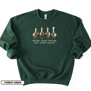 May include: Forest green sweatshirt with a graphic of four geese baristas, each holding coffee cups. The text "GRIND YOUR COFFEE. NOT YOUR TEETH." is printed below. The sweatshirt has a crew neck and long sleeves. The colour is described as forest green.