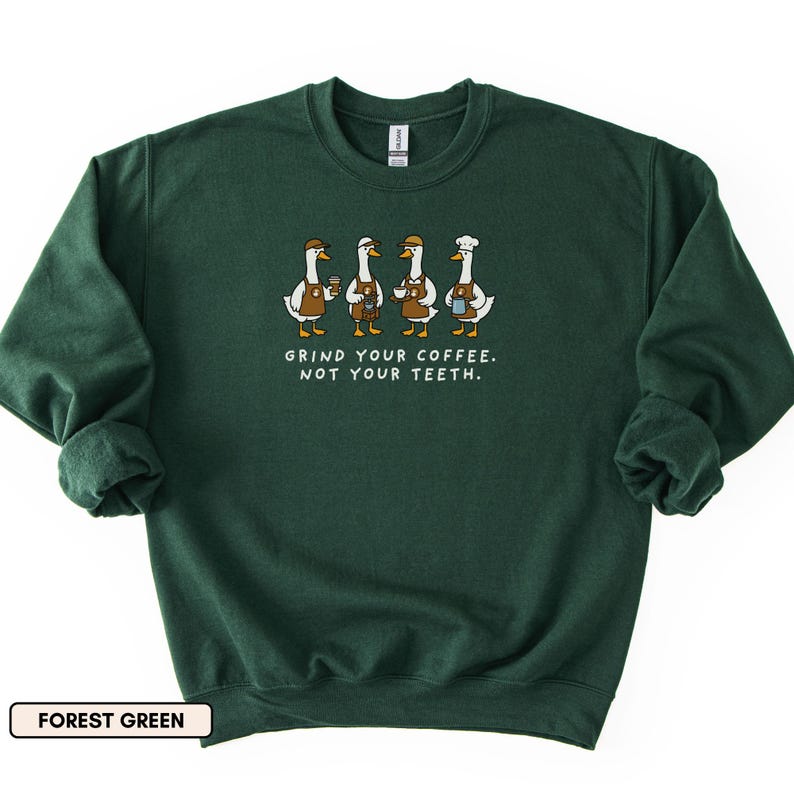 May include: Forest green sweatshirt with a graphic of four geese baristas, each holding coffee cups. The text "GRIND YOUR COFFEE. NOT YOUR TEETH." is printed below. The sweatshirt has a crew neck and long sleeves. The colour is described as forest green.