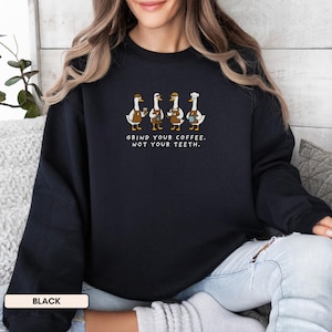 May include: Black sweatshirt with a fun illustration of four geese in barista outfits, each with coffee accessories. The text reads "GRIND YOUR COFFEE. NOT YOUR TEETH." The sweatshirt is a relaxed fit, perfect for everyday wear.