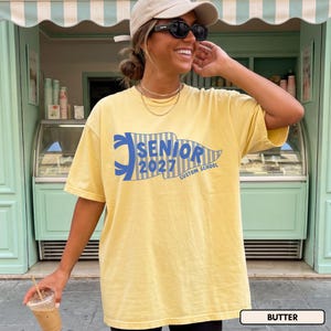 May include: A pale yellow short-sleeve t-shirt featuring a blue pennant graphic with vertical stripes and the text "SENIOR 2027 CUSTOM SCHOOL" printed in blue. The shirt has a crew neckline and a relaxed fit, showcasing a casual style.