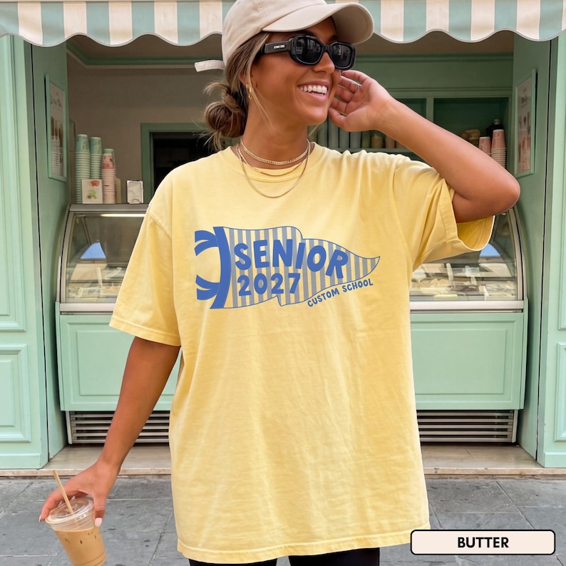 May include: A pale yellow short-sleeve t-shirt featuring a blue pennant graphic with vertical stripes and the text "SENIOR 2027 CUSTOM SCHOOL" printed in blue. The shirt has a crew neckline and a relaxed fit, showcasing a casual style.