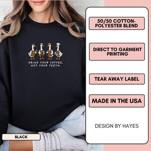 May include: Black sweatshirt featuring a design of four geese in aprons and hats, holding coffee cups. The text reads "GRIND YOUR COFFEE. NOT YOUR TEETH." It's made from a 50/50 cotton-polyester blend and printed directly onto the garment.