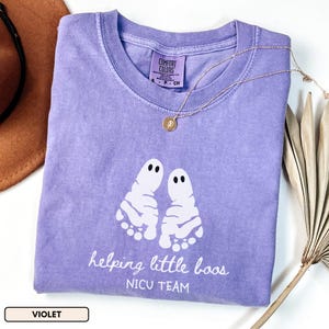 May include: A violet short-sleeved t-shirt featuring a white graphic of two ghost-shaped baby footprints. The text reads helping little boos NICU TEAM. The shirt has a crew neckline and a Comfort Colors brand label printed on the inside collar.