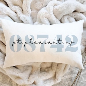 May include: A rectangular white pillow with the text "pt. pleasant, nj" in a light blue font. The pillow is resting on a soft, white, textured blanket. The numbers "08742" are also in light blue.