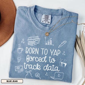 May include: A blue jean colored t-shirt with the text "BORN TO YAP forced to track data" in white. The shirt has white graphic icons of books, a coffee cup, a lightbulb, a computer, a bow, a magnifying glass, a laptop, a pen, and a graph.