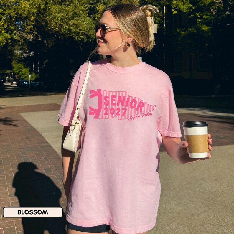 May include: A light pink short-sleeved t-shirt featuring a dark pink graphic of a pennant flag with the text SENIOR 2027 CUSTOM SCHOOL. The shirt has a crew neckline and a relaxed fit, worn with a white shoulder bag and dark sunglasses.