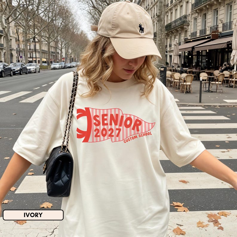 May include: An ivory t-shirt featuring a red and white striped pennant graphic with the text SENIOR 2027 CUSTOM SCHOOL, paired with a beige baseball cap with a small dark logo and a black shoulder bag with a gold chain strap.