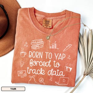 May include: A coral-colored t-shirt with the text "BORN TO YAP forced to track data" in white. The shirt features white graphic icons of books, a coffee cup, a lightbulb, and a laptop. A gold necklace with a pendant rests on the shirt.
