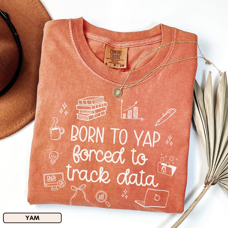 May include: A coral-colored t-shirt with the text "BORN TO YAP forced to track data" in white. The shirt features white graphic icons of books, a coffee cup, a lightbulb, and a laptop. A gold necklace with a pendant rests on the shirt.