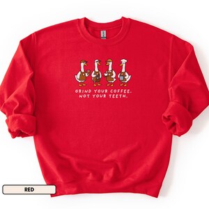 May include: A red crew neck sweatshirt. The front displays a graphic of four geese dressed as baristas, holding coffee cups and wearing aprons and hats. The text "GRIND YOUR COFFEE. NOT YOUR TEETH." is below the geese.